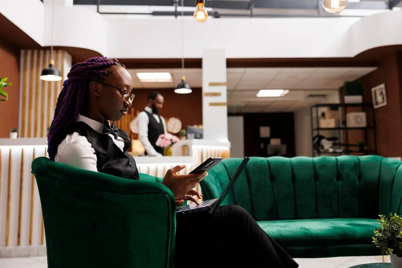 A young African American woman hotel manager sits in the reception lounge area, using a smartphone and a laptop to manage the budget and order supplies. Technology in the hospitality industry.